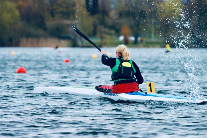 A woman seen from behind, paddling a kayak hard across a lake