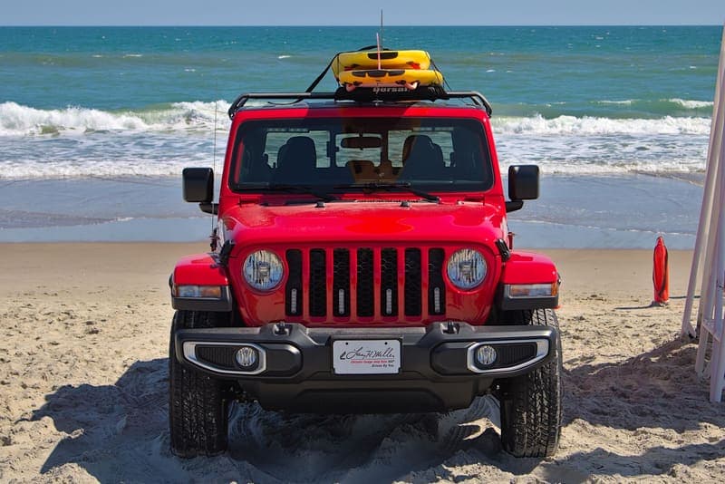 A tall red Jeep parked on a beach with a yellow boat strapped to its roof rack